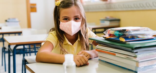 girl_sitting_at_her_chair_and_table_in_the_classroom_wearing_a_mask_to_protect_herself_during_the...