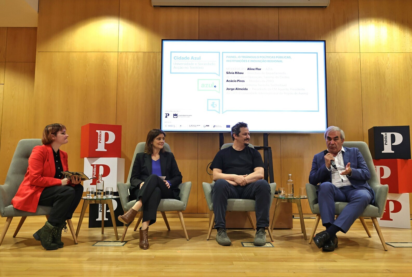 Painel de discussão com quatro participantes sentados em palco, com ecrã de apresentação ao fundo...